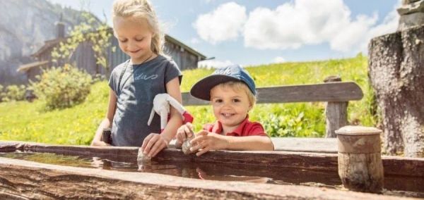 Dzieci bawiące się przy drewnianej rynnie ze źródlaną wodą symbolizują rodzinne lato na halach w Salzburskim Saalachtalu